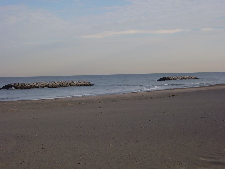 Beach with sand in the foreground, ocean with waves, and two rock breakwaters in the water under a cloudy sky
