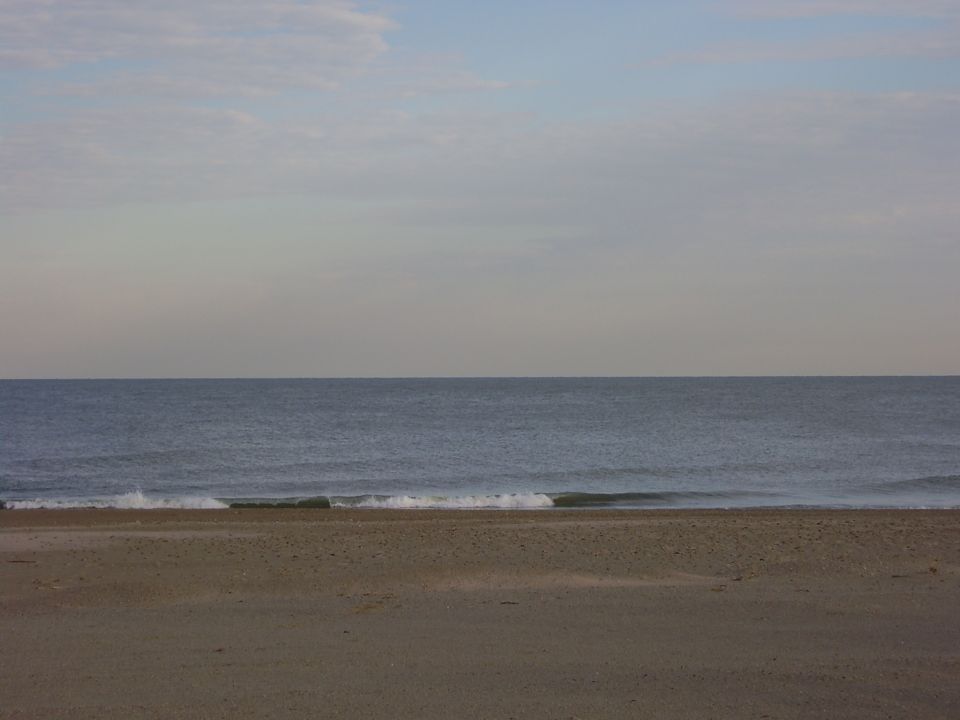 Beach with sand in the foreground and ocean under a cloudy sky. Watermark reads SUPERMOTORS