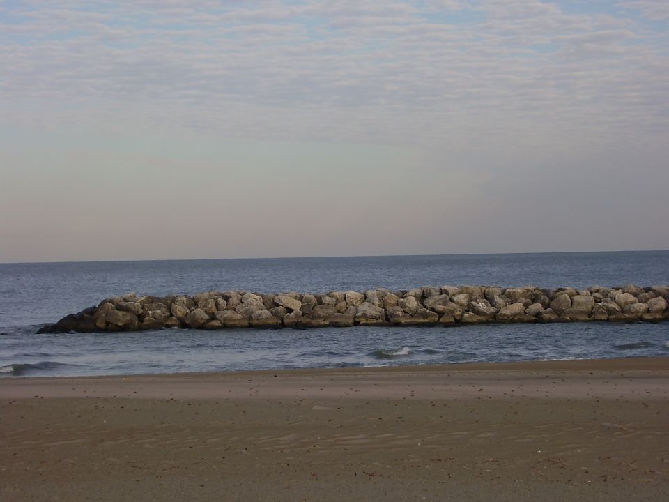 Beach scene with a rock breakwater and ocean under a cloudy sky. Supermotors.net logo in corner