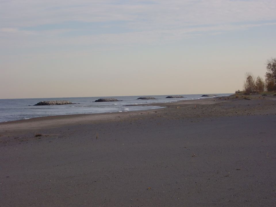 Beach with calm water, scattered rocks offshore, and a sandy shore under a hazy sky. Watermark reads SUPERMOTORS