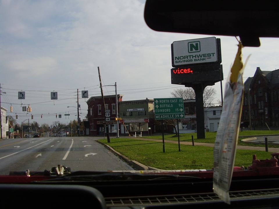 Street view from inside a vehicle, showing a bank sign, directional signs, and buildings under a cloudy sky