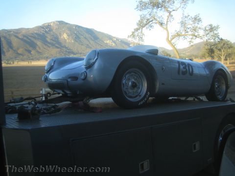 1990 Ford Bronco - silver Porsche 550-style spyder race car numbered 130 rides on a flatbed truck with mountains in the background.
