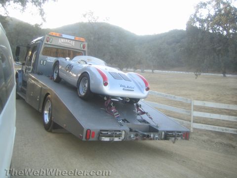 1990 Ford Bronco - silver-and-red spyder race car sits chained on an American Transportation flatbed truck parked in a grassy field.