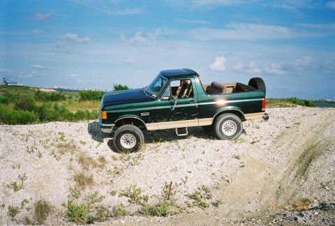 1990 Ford Bronco - A green Ford F-Series truck is parked on a dirt road with sparse vegetation under a blue sky.