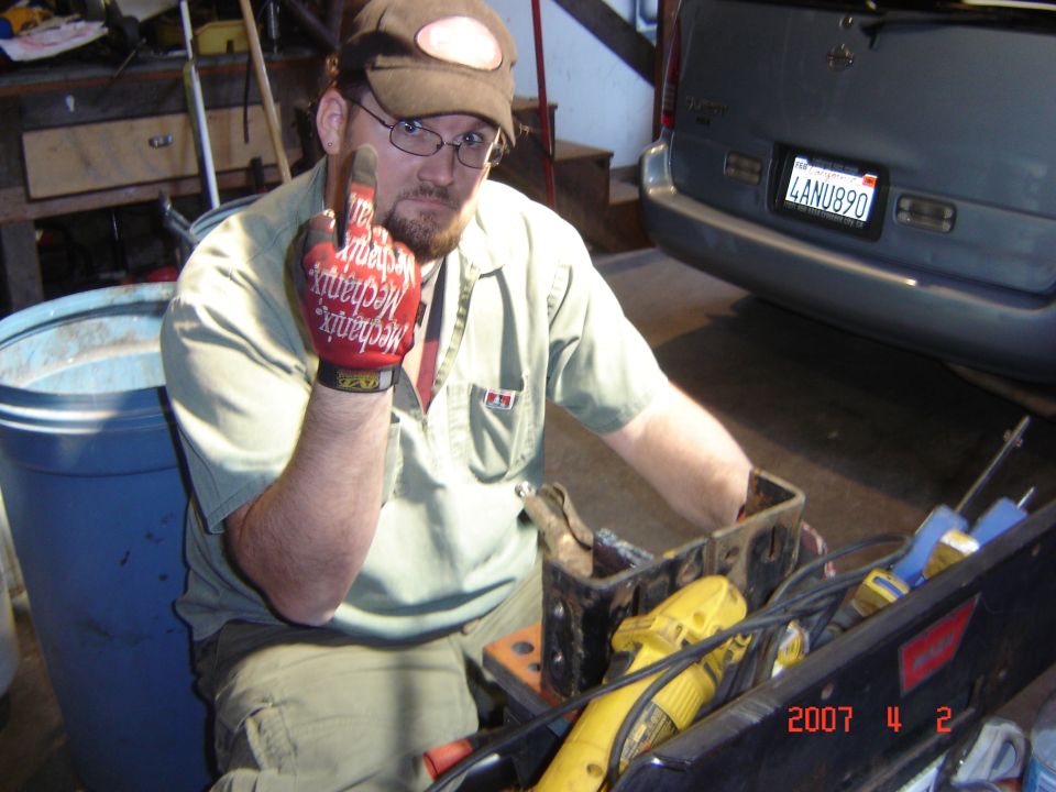 Man wearing a cap and mechanic's gloves gives the middle finger while sitting in a garage. A blue trash can is to his left, and a silver SUV is parked behind him