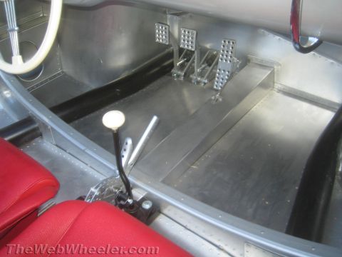 1990 Ford Bronco - Bare aluminum footwell of a silver vintage racing car with three drilled pedals, a white-knobbed shifter and a red seat edge.