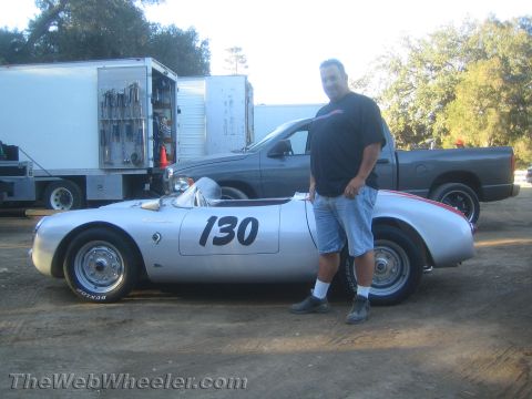 1990 Ford Bronco - man in a black shirt and denim shorts stands next to a silver vintage racing roadster numbered 130 at an outdoor paddock.