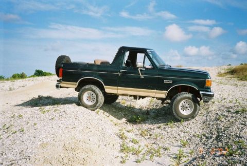 1990 Ford Bronco - This 1990 Ford Bronco is seen from a 3/4 front angle, parked on a dirt and gravel terrain under a cloudy sky.