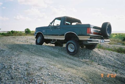 1990 Ford Bronco - This Ford Bronco is parked on a gravel incline with a body of water and vegetation in the background.