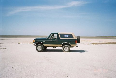 1990 Ford Bronco - A 1990 Ford Bronco is seen from the side, parked in a desolate, sandy area with a clear blue sky above.