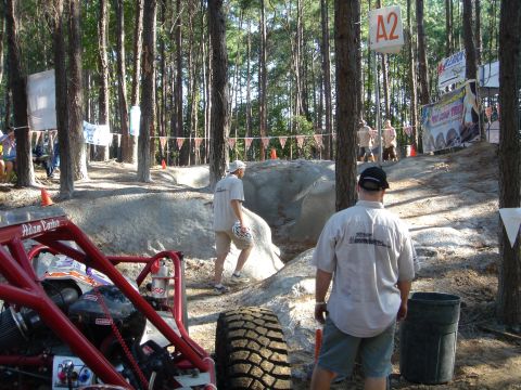 2002 Jeep Wrangler - A red off-road buggy with large tires is parked in a wooded area with spectators and an obstacle course in the background.