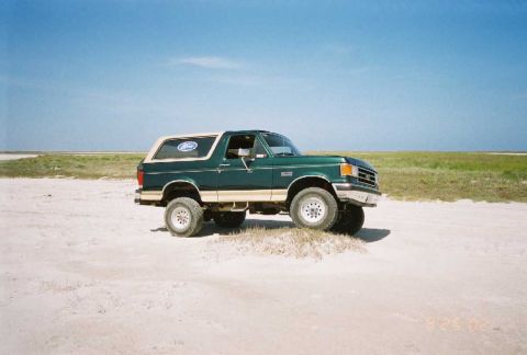 1990 Ford Bronco - This green 1990 Ford Bronco, with its distinctive tan lower body and white roof, is captured in a three-quarter front view on a sandy lands…