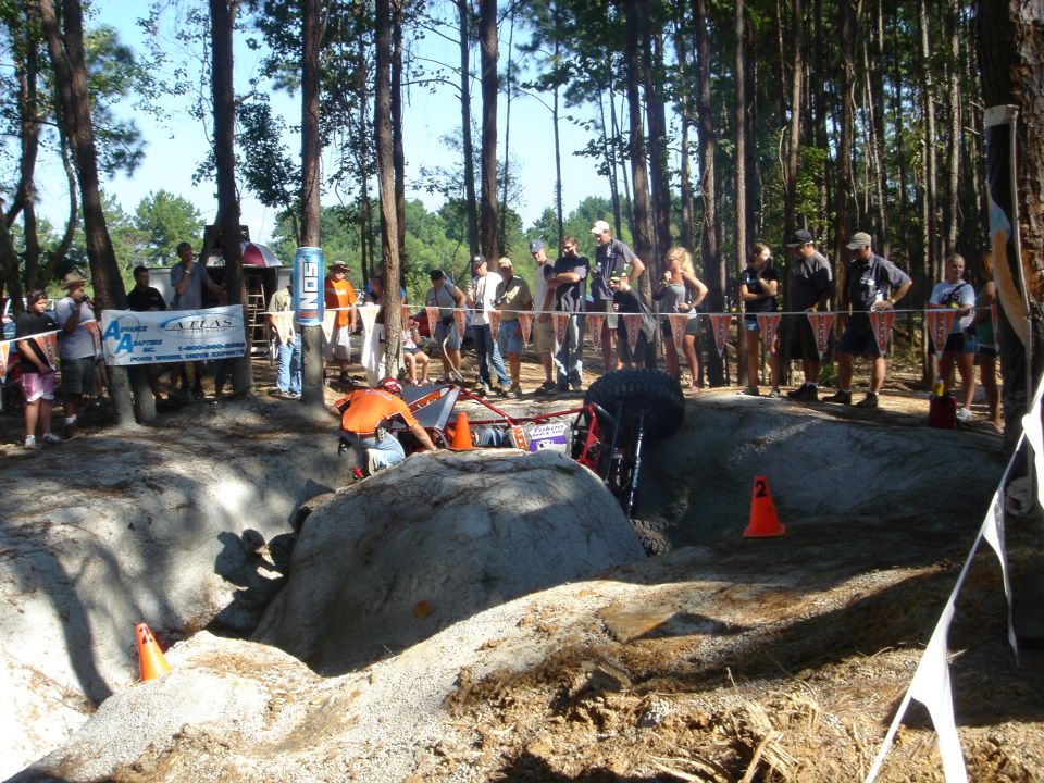 Orange off-road rig climbing concrete boulder obstacle at a 4x4 event with spectators