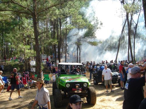 2002 Jeep Wrangler - A green Jeep Wrangler is parked in a wooded area with spectators around. Smoke billows in the background.