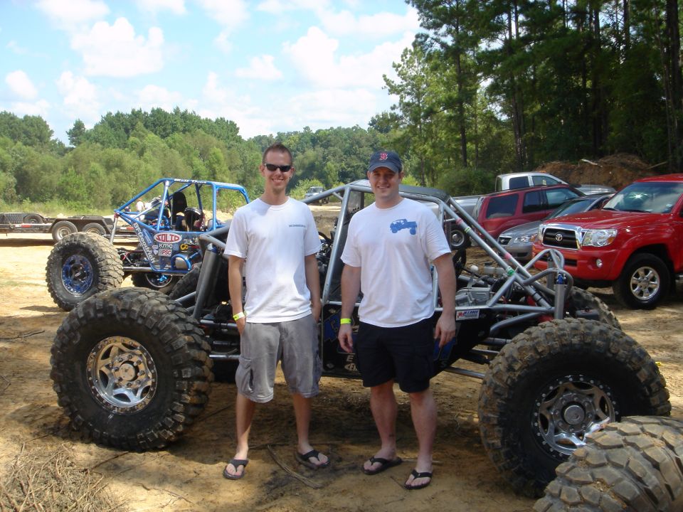 Two men standing beside a silver tube-chassis rock crawler buggy at an off-road park