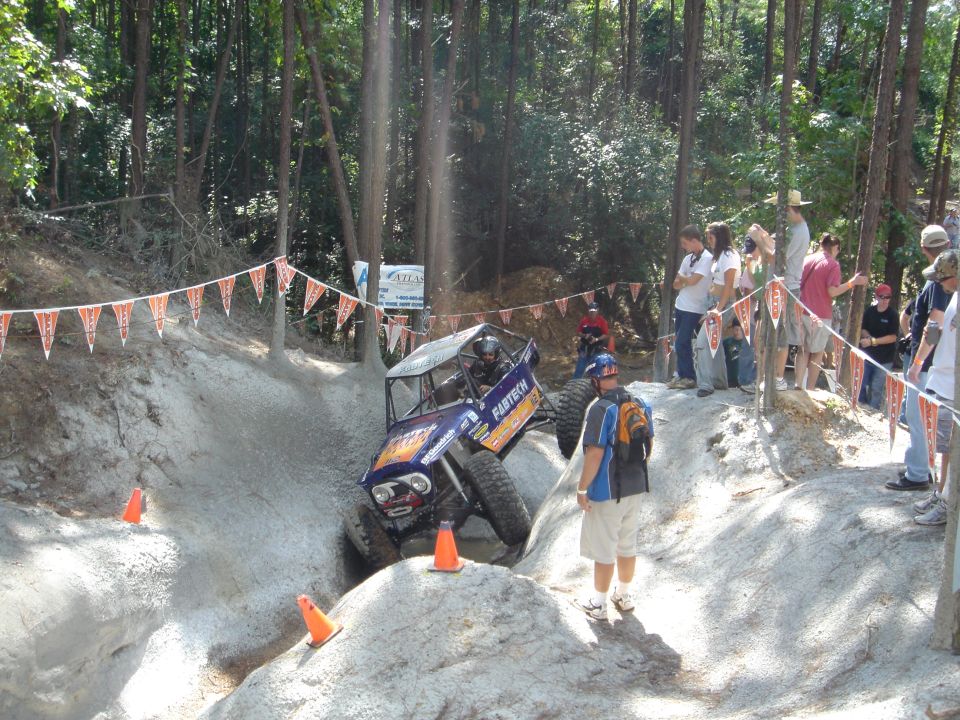 Purple tube-chassis rock buggy on a sandy competition obstacle with spectators watching.