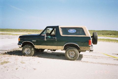 1990 Ford Bronco - A 1990 Ford Bronco with a tan top and spare tire mounted on the rear is seen on a sandy terrain under a clear blue sky.