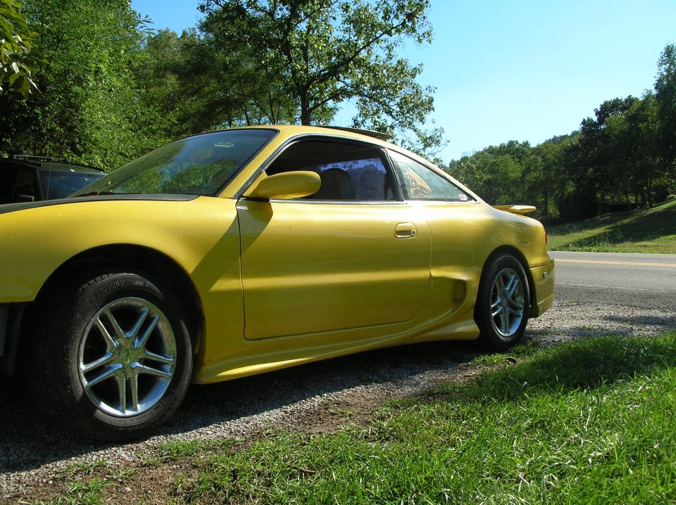 Yellow 1993 Mazda MX-6 parked on a gravel shoulder next to a road, side view