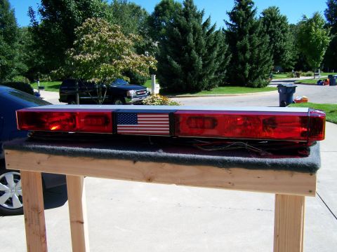 2003 Ford Crown Victoria - An emergency light bar with an American flag decal is displayed on a wooden stand outdoors.