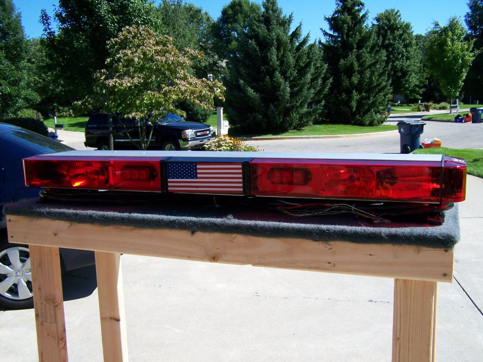 Emergency light bar with American flag decal, sitting on a wooden stand. A black pickup truck is visible in the background
