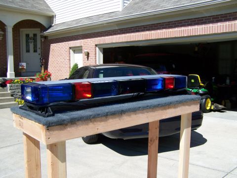 2003 Ford Crown Victoria - A police light bar is displayed on a stand, with a dark sedan and a riding mower visible in the background.