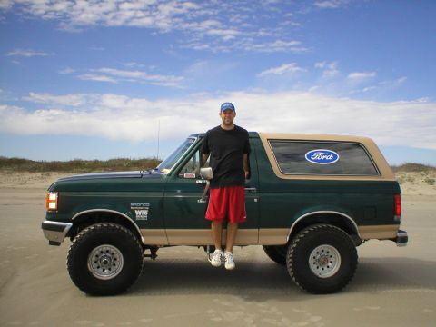 1990 Ford Bronco - A man stands beside a two-tone Ford Bronco parked on a sandy beach under a cloudy sky.