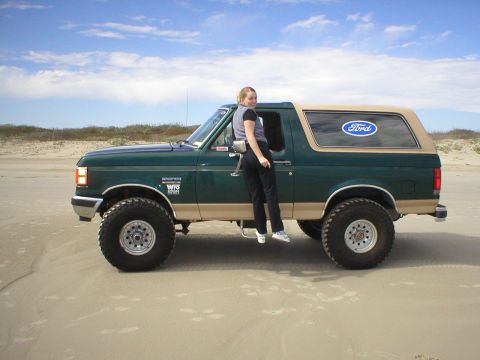 1990 Ford Bronco - A woman poses with a green 1990 Ford Bronco on a sandy beach under a partly cloudy sky.