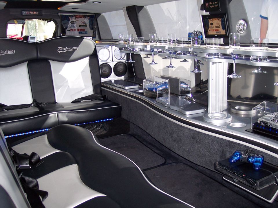 Limousine interior showing seats, bar with glasses, screens, and a game console