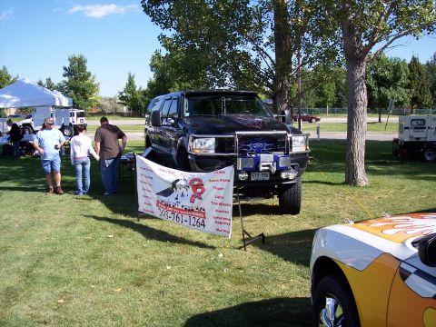 1972 Ford Ranchero - A modified black Ford truck is displayed at an outdoor event, with a banner advertising accessories and a phone number.