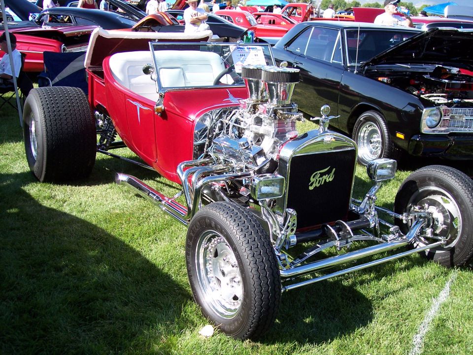Red hot rod with Ford grille and exposed V8 engine. Black car in background