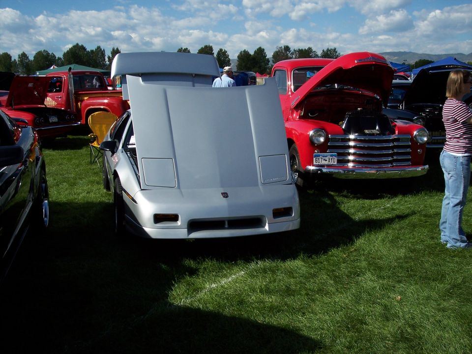 Silver sports car with hood open, parked on grass next to a red pickup truck