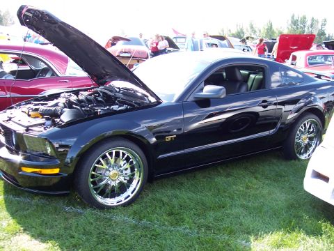1972 Ford Ranchero - A black Ford Mustang GT is displayed with its hood open, revealing the engine and chrome wheels at an outdoor car show.