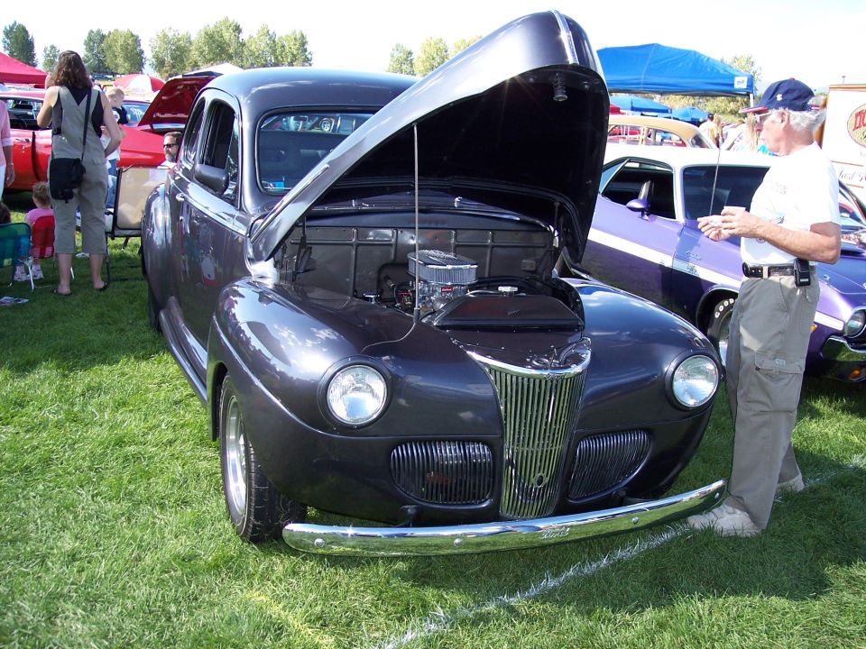 dark gray coupe with hood open, chrome grille, and a man standing next to it. Another purple car is visible behind