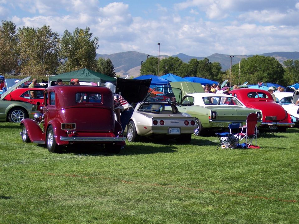 Several classic cars parked on grass at an outdoor show, with tents and mountains in the background