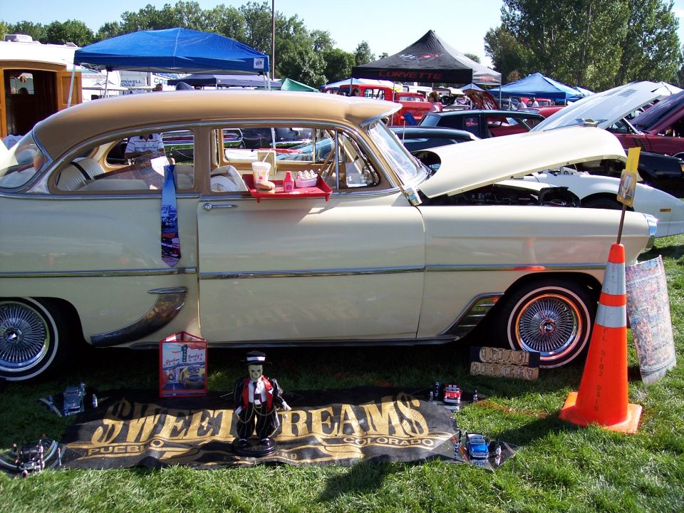 Cream-colored 1954 sedan with a tan roof, chrome trim, and wire wheels, parked on grass with a food tray on the door