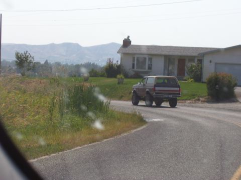 1990 Ford Bronco - A Ford Bronco drives away from the camera on a country road, with a house and mountains in the background.