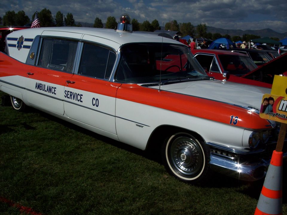 Side view of a white and orange ambulance with 'AMBULANCE SERVICE CO' text. Another car is parked nearby