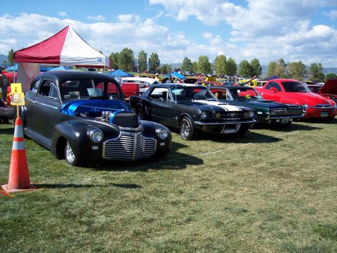 1972 Ford Ranchero - A row of classic cars are on display at an outdoor event, with a dark gray sedan with its hood open in the foreground.