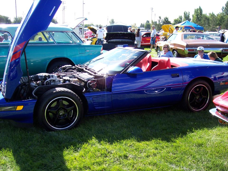 Blue convertible with hood open showing engine, red interior, parked on grass at car show with other vehicles