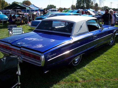 1972 Ford Ranchero - A blue 1965 Ford Thunderbird with a white vinyl roof is displayed at a car show with other vehicles and tents in the background.