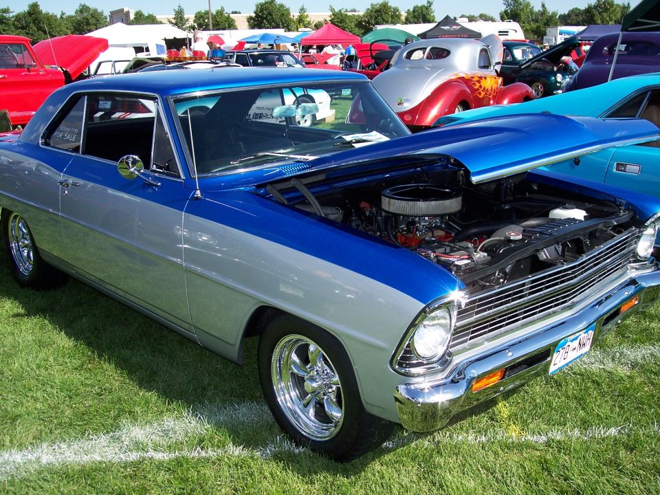Blue and silver coupe with hood open, showing engine. Other cars and tents in background