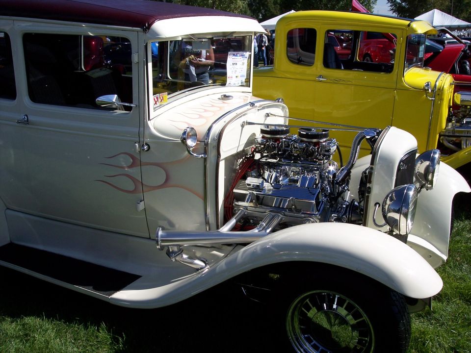 White hot rod with flames and exposed chrome engine, parked on grass next to a yellow car