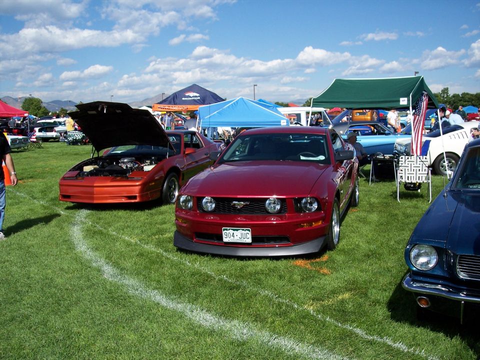 Red 2005 Ford Mustang with license plate 904-JUC parked on grass next to an orange Pontiac Firebird with hood open