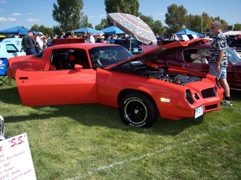 1972 Ford Ranchero - A red sports coupe is displayed with its hood and door open at a car show, with other vehicles and tents in the background.
