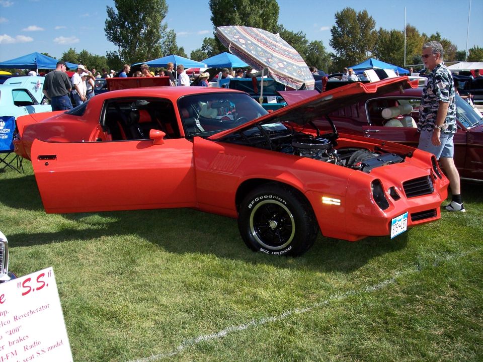 Red sports coupe with hood open and driver door ajar, parked on grass at an outdoor car show