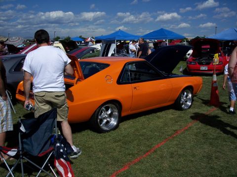 1972 Ford Ranchero - Orange AMC AMX parked on grass at an outdoor event with other vehicles and tents in the background.