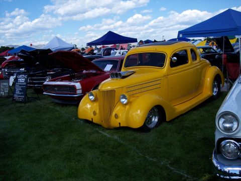 1972 Ford Ranchero - A bright yellow hot rod coupe is displayed at an outdoor car show, with other vehicles and tents in the background.