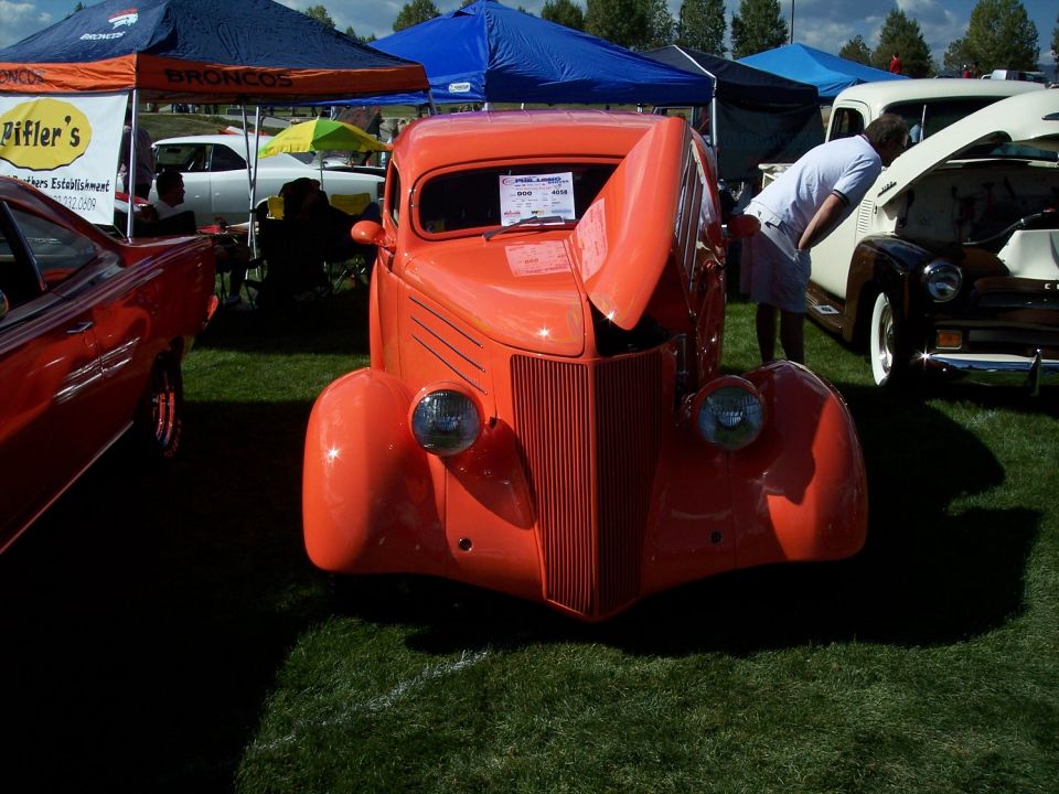 Orange hot rod coupe with hood open, parked on grass at an outdoor car show