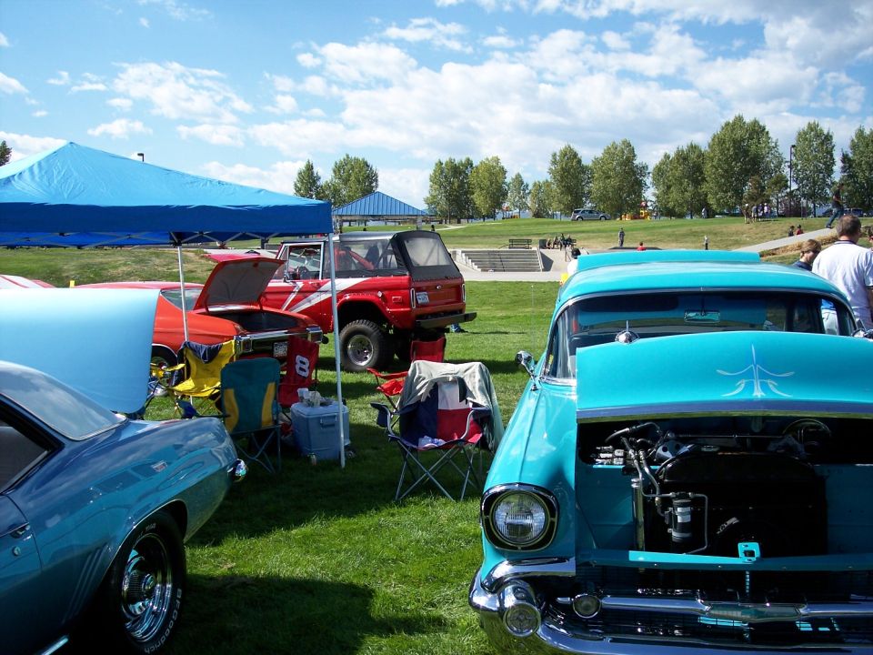 Several classic cars, including a teal 1950s model with hood open, are parked on grass under a blue tent and a clear sky