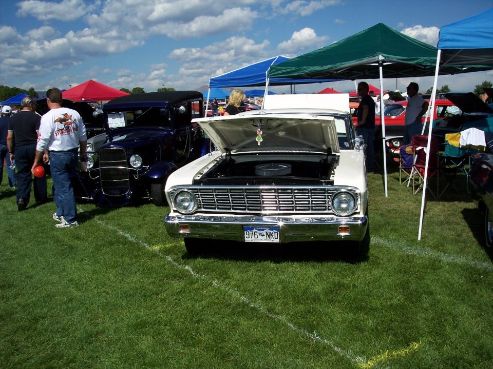 White 1964 Ford Falcon with hood open, parked on grass at car show. Blue 1930s hot rod in background